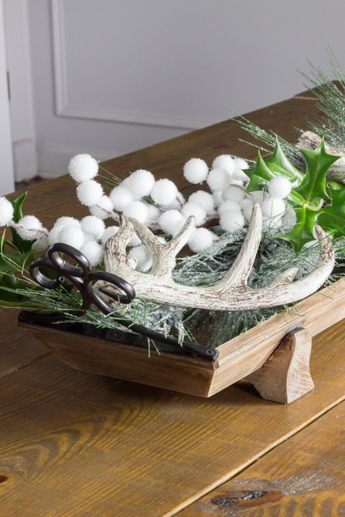 A wooden sleigh tray of frosted greenery, holly leaves, white snowball berries and a faux antler on a farmhouse table